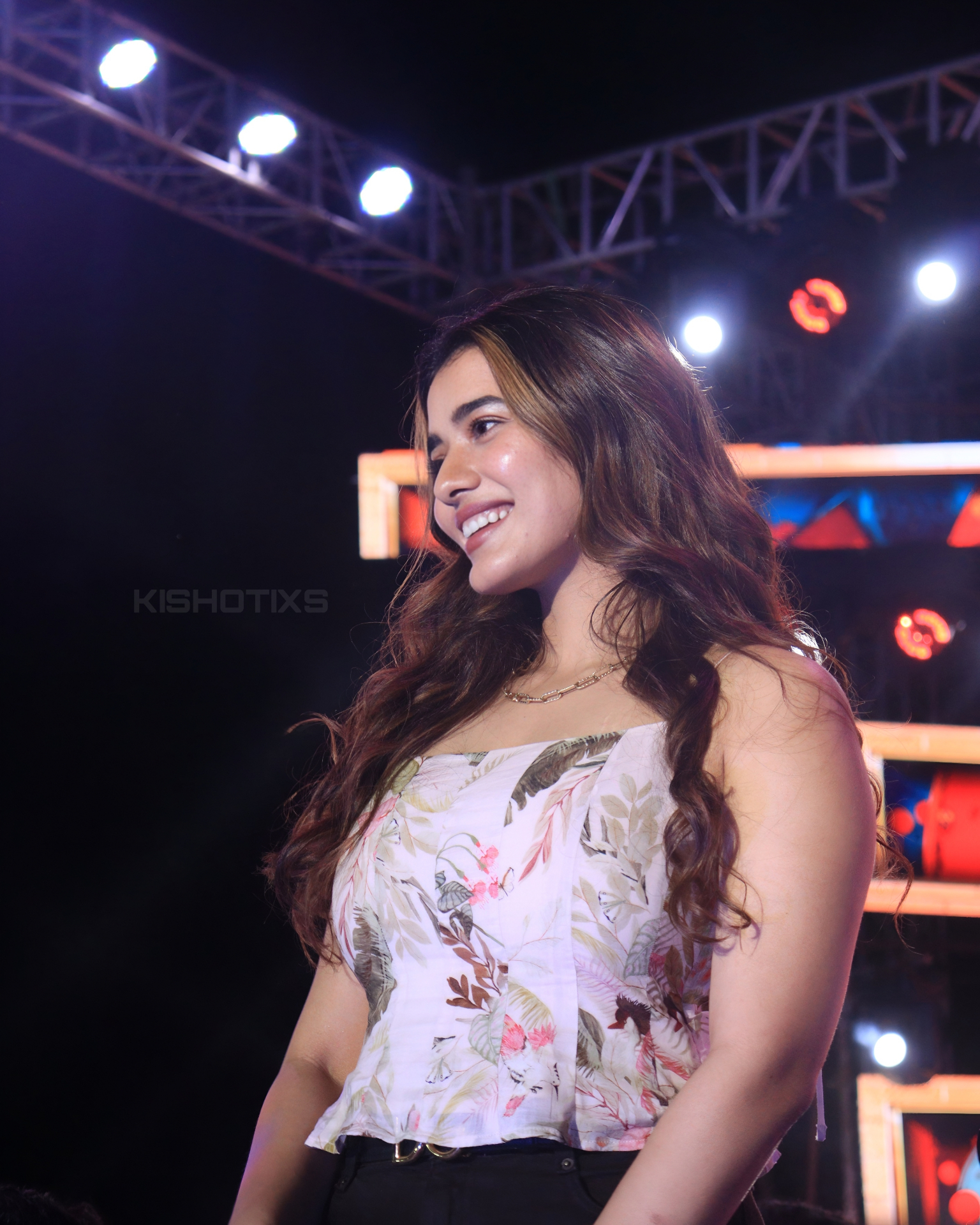 Smiling long-haired performer in a floral corset top looking off-camera under stage truss lighting