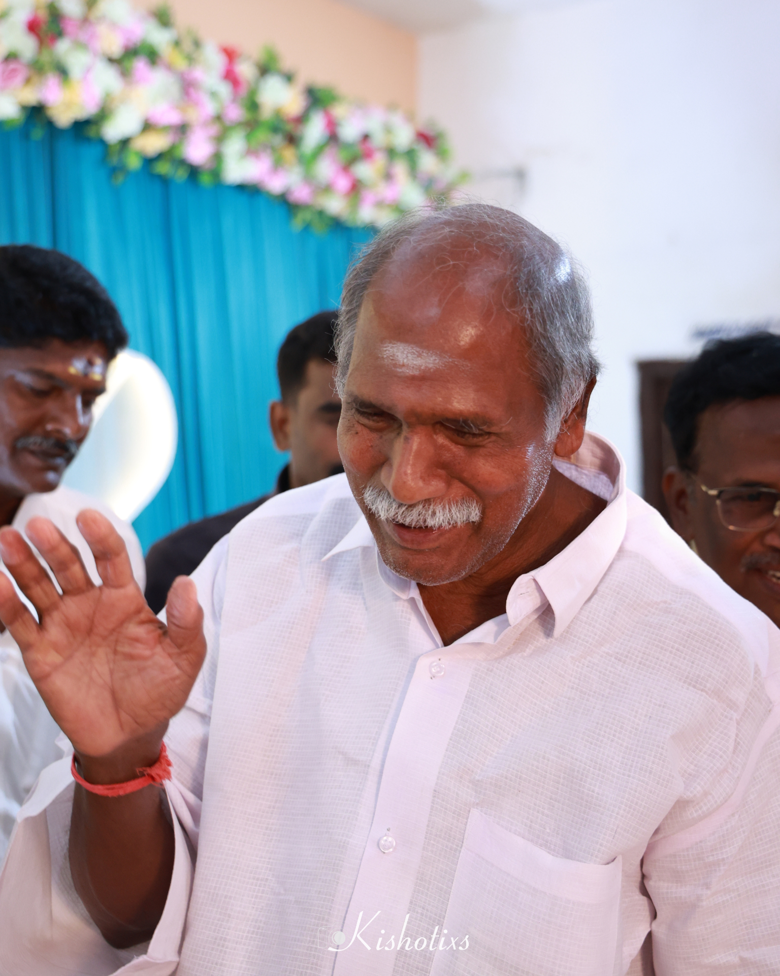 Elder gentleman in a pale pink shirt raising his hand in blessing at a family celebration