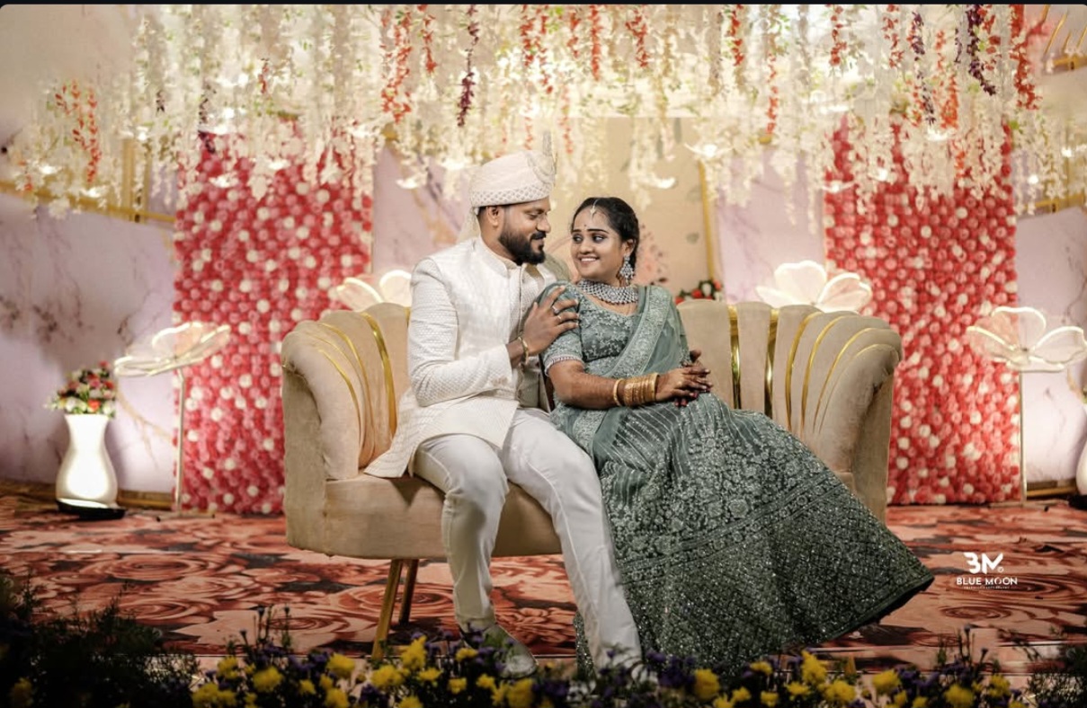 Groom in white sherwani and bride in green lehenga posing on a plush sofa in front of a red and white floral reception backdrop