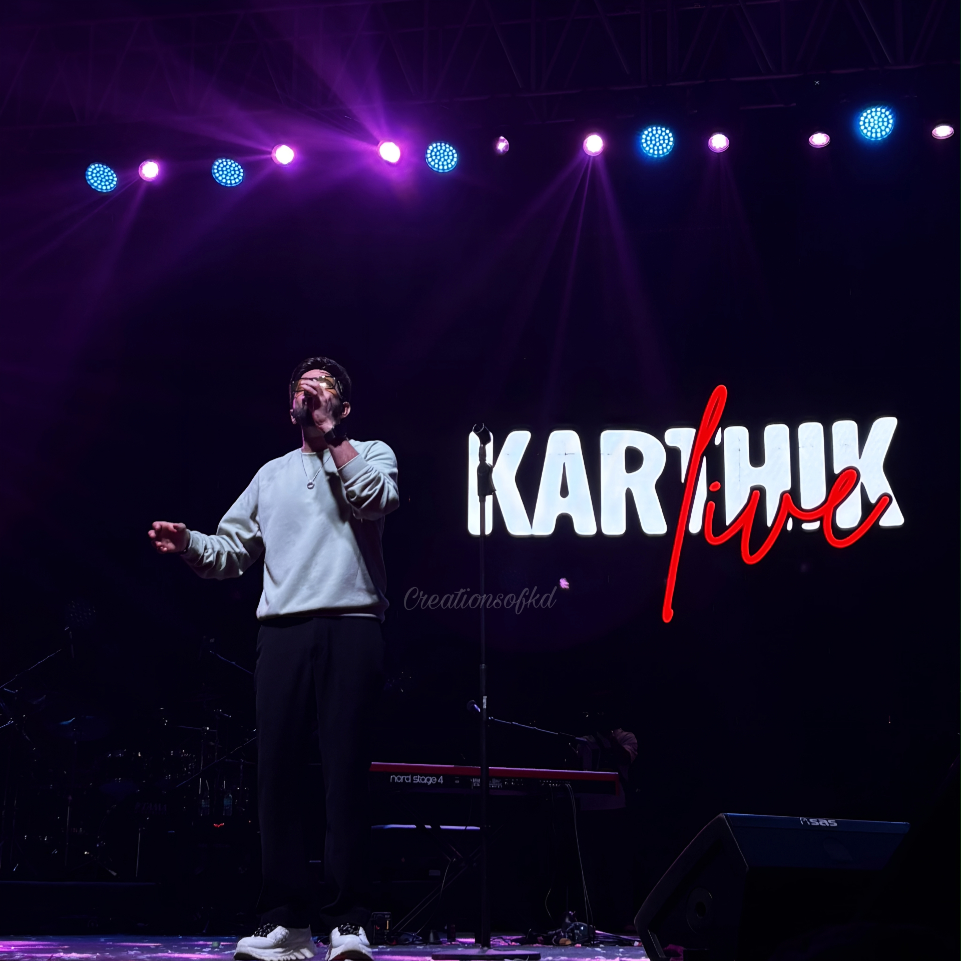 Male singer performing on stage in front of a Karthik Live LED sign under purple and blue stage lights